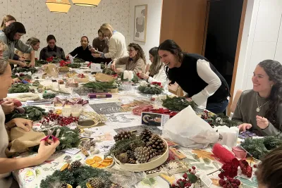 Ambiente animado en un taller de Navidad, con un grupo de mujeres riendo y trabajando en sus coronas de Adviento con piñas, lazos y ramas.