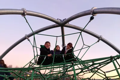Tres niñas sentadas en la cima de una cúpula de cuerdas de un parque bajo un cielo despejado al atardecer.