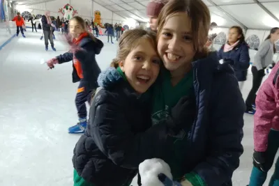 Dos niñas con abrigos oscuros y guantes se abrazan y sonríen a la cámara en la pista de hielo, una de ellas sosteniendo un poco de nieve.