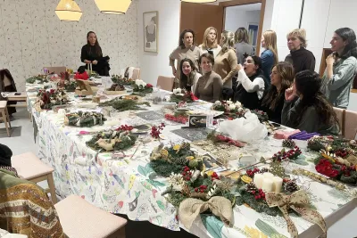 Grupo de mujeres interactuando y hablando en un ambiente social, rodeando una mesa larga con varias coronas de Adviento ya terminadas.