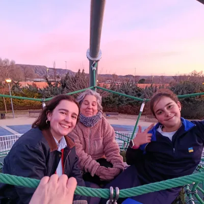 Dos jóvenes y una mujer mayor sonriendo sentadas en una red de cuerdas de un parque durante el atardecer.