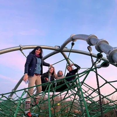 Cuatro personas posando en lo alto de una estructura piramidal de cuerdas en un parque infantil con cielo rosado.