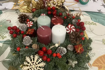 Corona de Adviento tradicional con cuatro velas de colores (verde claro, rojo, burdeos y blanco) rodeadas de ramas de abeto, piñas, bayas rojas y adornos navideños.