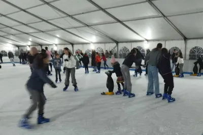 Escena de la pista de hielo cubierta, varias personas patinando, incluyendo una niña pequeña que usa un patín auxiliar con forma de pingüino.