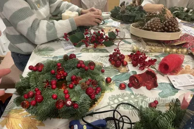 Primer plano de una corona de Adviento decorada con muchas bayas rojas y una persona trabajando al fondo, con una pistola de silicona sobre la mesa.