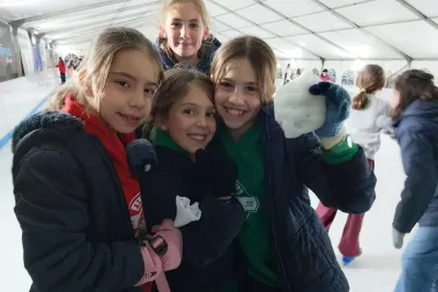Primer plano de cuatro niñas sonriendo juntas en la pista de hielo, vestidas con ropa de invierno.