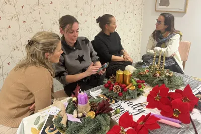 Cuatro mujeres conversando animadamente alrededor de una mesa decorada con dos coronas de Adviento terminadas, una morada y otra dorada con piñas.